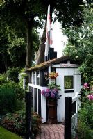 Quaint painted garden shed with thatched roof and flag, stone sett path and gate, reclaimed chimney pot with Hydrangea, adjacent dense planting with mature trees, shrubs, Buddleia and Phlox - 'Trevinia', Stubbins, Lancashire, NGS
