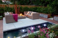 Small garden lit up at night, with wicker sofas on decked and paved patio, backed by Fargesia murielae - Bamboo hedge. Rectangular pond with row of square water features and lights 
 
