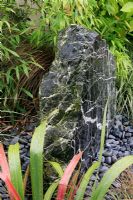 Drilled marbled rock discharging water through a bed of pebbles into an underground sump and surrounded with grassy leaved foliage including bamboo, Phormium and Carex