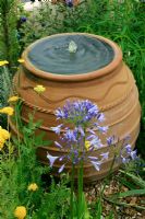 Cretan terracotta oil jar fountain discharging water into a gravel bed with Agapanthus and Achillea in the foreground