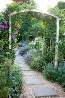Gravel path under wooden arch with Clematis growing up it in country garden.
