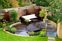 Overview of small urban garden in September with contemporary furniture and patio heater on circular sunken decked terrace. Raised bed and planting of Phormium, Arbutus unedo tree and Lavandula. Muswell Hill, London