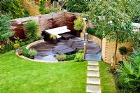 Overview of small urban garden in September with contemporary furniture and patio heater on circular sunken decked terrace. Raised bed and planting of Phormium, Arbutus unedo tree and Lavandula. Paving slab path across lawn. Muswell Hill, London