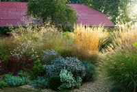 Central circular bed is a mass of graceful grasses and herbaceous perennials including Miscanthus 'Morning Light', Stipa gigantea, Calamagrostis x acutiflora 'Karl Foerster', purple sage and Sedum 'Purple Emperor' with striking red tin roof on barn behind - Grass Garden, Hants