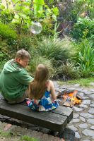 Man and child sitting on wooden bench by firepit on circular cobblestone patio in urban garden. Glass hanging sound speaker above. Yulia Badian, London, UK