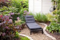 Small gravel patio with lounger surrounded by planting of Corylus avellana 'Purpurea' and Alliums