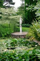 Classical statue on a tall plinth set on a raised circle at the edge of a Nymphaea - Waterlily pond. Cornus alternifolia 'Argentea' -  Pagoda or Wedding Cake tree behind. Sheephouse, Painswick