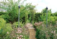 Path leading through finial topped posts, framing the view to metal arbour and pot beyond. Fovant Hut Garden, Wilts