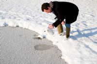 Man making hole in ice of wildlife pond by pouring warm water from kettle. Snow surrounding.