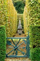Blue gate leading to clipped Fagus and Taxus baccata hedges and stone obelisk - Silverstone Farm, Norfolk
