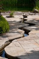 A meandering canal leads through a flagstone paving towards a beach with pebbles that belongs to a natural swimming pool. 
