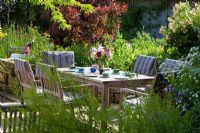 Wooden garden furniture with blue striped linen cushions in a slightly shaded area in June. Planting includes Rosa 'Ghislaine de Féligonde', Cotinus coggygria 'Royal Purple' and Tanacetum parthenium 