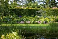 View over a natural swimming pool to a pavillon, backed with a hedge. The slope is secured by lime sand boulders, planting includes Alchemilla mollis, Buxus, Carpinus betulus, Fagus, Geranium, Hosta, Larix, Lupinus and Salix