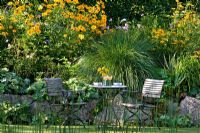 Wooden garden furniture in front of a raised perennial border which is secured with lime sand boulders and planted with Alchemilla mollis, Heliopsis, Miscanthus sinensis and Rudbeckia fulgida