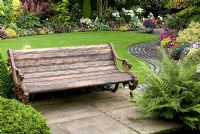 Old wooden bench in secluded, colourful garden with dense plantings of shrubs, conifers, ferns and bedding plants, path made from reclaimed slabs and cobbles - Brocklebank Road, Southport, Lancashire NGS
