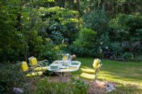 Outside dining area with white wood and iron garden furniture with yellow cushions. Rosa 'Mary Rose', Digitalis purpurea, Heuchera micrantha 'Palace Purple',  Ligustrum and Viburnum plicatum in borders