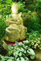 Chinese buddha surrounded by foliage plants including Brunnera, Choisya and Fatsia. Beggars Knoll, Newtown, Westbury, Wiltshire, UK