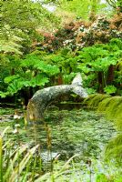 Whale's tail sculpture in the pond with Gunnera manicata behind and flowering Pieris beyond - Trewidden, Buryas Bridge, Penzance, Cornwall, UK