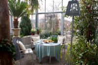 Inside the conservatory, a rest area with a view to the outside terrace. A table is laid for breakfast, wood and metal chairs, linen cushions and a modern styled lantern contrasts with classical terracotta pots - Wintergarten, Germany