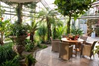 View into the glasshouse with tropical climate, wicker chairs and wooden table with a fruit arrangement. Planting includes Anthurium andreanum, Araucaria heterophylla, Beaucarnea recurvata, Billbergia nutans, Citrus sinensis, Cycas revoluta and Phoenix roebelinii - Wintergarten, Germany