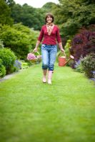 Woman wearing blue jeans and wellies carrying a red watering can and a wooden trug of pink chrysanthemum