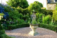 Formal garden with central urn on gravel path is planted with yellow and blues including Rosa 'Agnes', Allium cristophii and Lupinus. Path lined with Nepeta 'Six Hills Giant' - Catmint, leads toward white dovecot and bed of Irises - Old Rectory, Kingston, Isle of Wight, Hants, UK