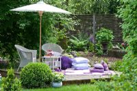 Wooden deck with white wicker garden furniture, parasol and a blanket with white and lilac cushions. Plants are Buxus, Fern, Hydrangea macrophylla and Lupinus