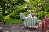 Patio with table set for meal, wooden chairs, and a rusty metal chandelier next to planting of Buxus, Paeonia and Rosa