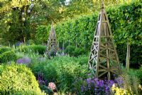 Summer border with wooden obelisk, backed by pleached Tilia - Lime hedge. Plants include Allium christophii, Penstemon, Achemilla mollis, Hesperis
