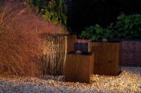 Wooden cube water features in gravel next to low brick wall, lit up at night. Epimedium x versicolor 'Sulphureum', Anemanthele lessoniana
