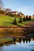 Plas Cdanant Gardens, Menai Bridge, Anglesey, Wales. April. Tank in the Walled Garden. Bergenia 'Overture' at edge. Yew pyramids. 