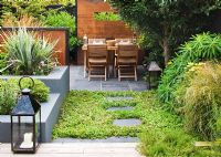 Small town garden with wooden table and chairs, lantern, screens made of mild steel, and raised beds with lush planting, London 
