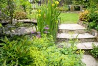Stone path and steps leading to raised circular stone edged pond with adjacent lawn and perennials 