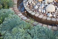 A circular rill of water between planting of silver foliage grasses, perennials, gravel, pebbles and brick 