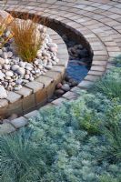 A circular rill of water between planting of silver foliage grasses and perennials, gravel, pebbles and brick in the 
