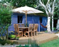 Blue summerhouse with decking, table, chairs and parasol, birch trees and hammock 