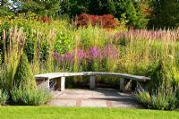 Curved wooden seat backed by grasses and flowering perennials including Lythrum virgatum 'Dropmore Puprle' along the main borders -  RHS Garden Harlow Carr, Harrogate, North Yorkshire, UK
