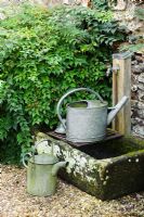Watering cans beside outdoor tap mounted over old stone sink - Barnwells, Cerne Abbas, Dorset.