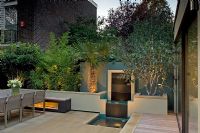 Urban courtyard garden lit up at dusk with decking, raised beds with Bamboo and water feature, London, UK
