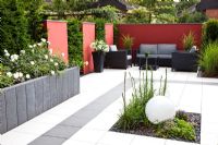 Modern paved garden with raised beds backed by red screen and Taxus - Yew hedge. Plants include Miscanthus and Rosa 'Schneeflocke'
