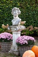 Chrysanthemum in baskets either side of a stone bust on a plinth - Huys en Hof