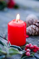 Lit red candle on frosty table with Holly and cones