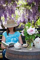 Woman reading a book in the shade under Wisteria, vase of Paeonia and Deutzia