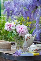 Vase of Paeonia and Deutzia with tea set, book and sun hat, Wisteria in background
