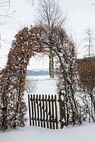 A hornbeam arch with a wooden gate separates a country garden from the landscape