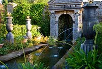 Rill pool flanked by turned oak urns spouting water through gilded lions' heads, with golden agaves in their tops. Planting includes Alchemilla mollis and white agapanthus. Water emerges from a rocky grotto framed by a scallop shell pediment and muscular caryatids. The Collector Earl's Garden designed by Julian and Isabel Bannerman.