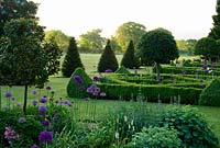 Parterre of box hedges containing standard bays, planted with santolina and Allium 'Purple Sensation' with yew pyramids and lawn stretching out toward a ha ha and Dorset countryside beyond. Old Rectory, Pulham, Dorset, UK