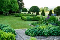 Alchemilla mollis seeded into terrace in foreground. Beyond is a parterre of box hedges containing standard bays, planted with santolina and Allium 'Purple Sensation' with yew pyramids and lawn stretching out toward a ha ha and Dorset countryside beyond. Old Rectory, Pulham, Dorset, UK
