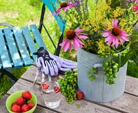 On a wooden table a vase with coneflower, Ladys Mantle and grapes. Also on the table is gardening gloves, hand fork, a bowl of strawberries and a glass of water. In the background a colourful turquoise garden chair.