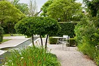 Bistro table and chairs on a gravel area in a formal designed garden with water canal, clipped shrubs and trees - Amelanchier, Anthemis tinctoria, Fagus sylvatica 'Purpurea', Iris sibirica and Malus - Germany